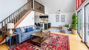 A spacious living room with a wooden staircase, a blue sofa, a coffee table, and a red patterned rug. - Forest 2BR chalet w/ hot tub & backyard (Gerrardstown)