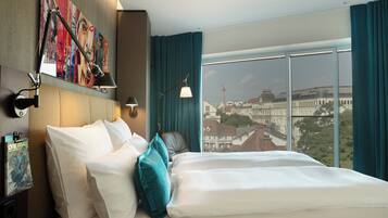 Ein Hotelzimmer mit einem großen Bett, einer Bettseite mit Kunstwerk, einem Schreibtisch mit Lampe und Blick auf eine Stadtansicht durch große Fenster.