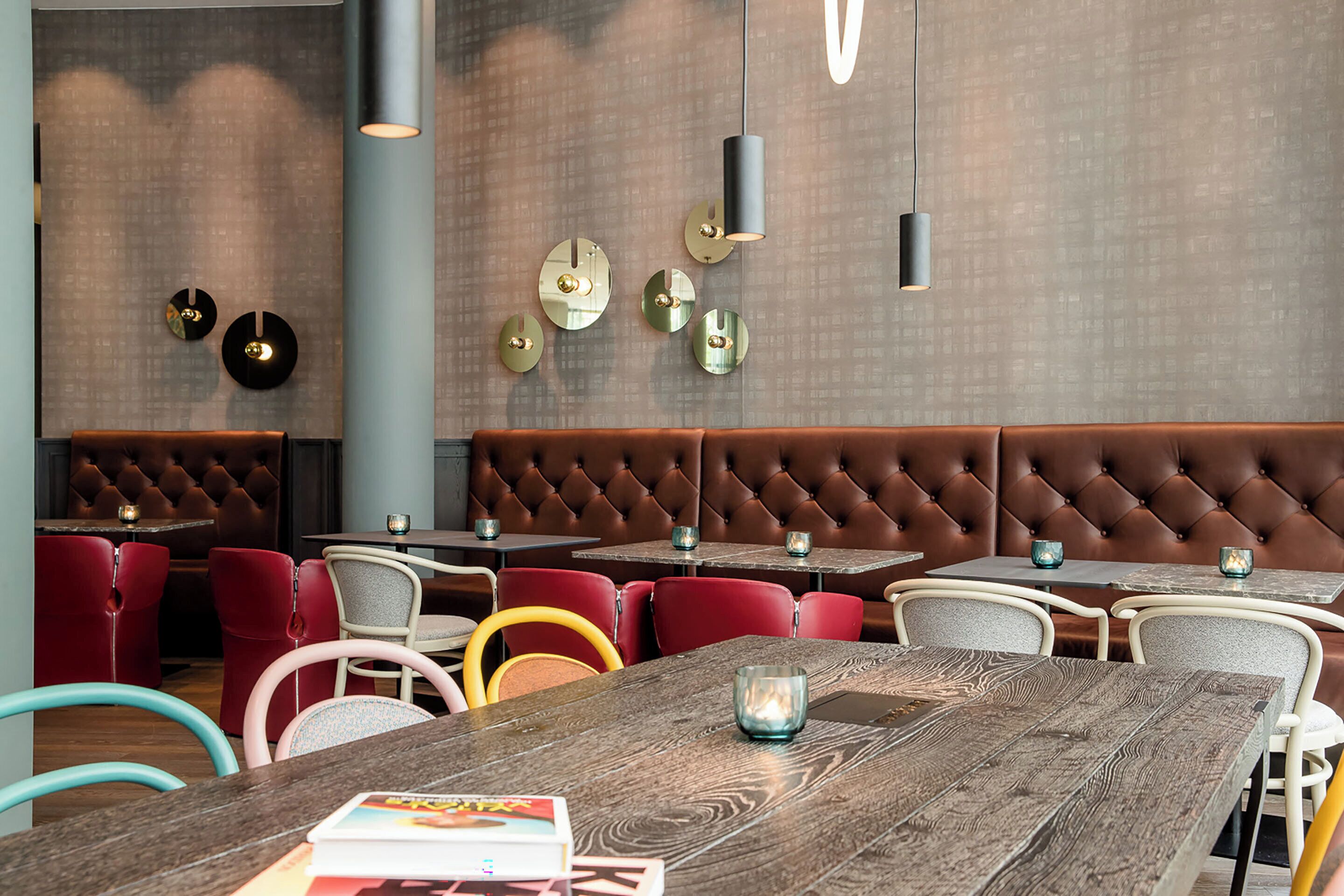 Un interior de restaurante moderno con un área de asientos tipo banca tapizada en tonos marrones, sillas rojas y blancas, y mesas de madera.