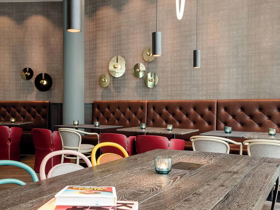 Un interior de restaurante moderno con un área de asientos tipo banca tapizada en tonos marrones, sillas rojas y blancas, y mesas de madera.