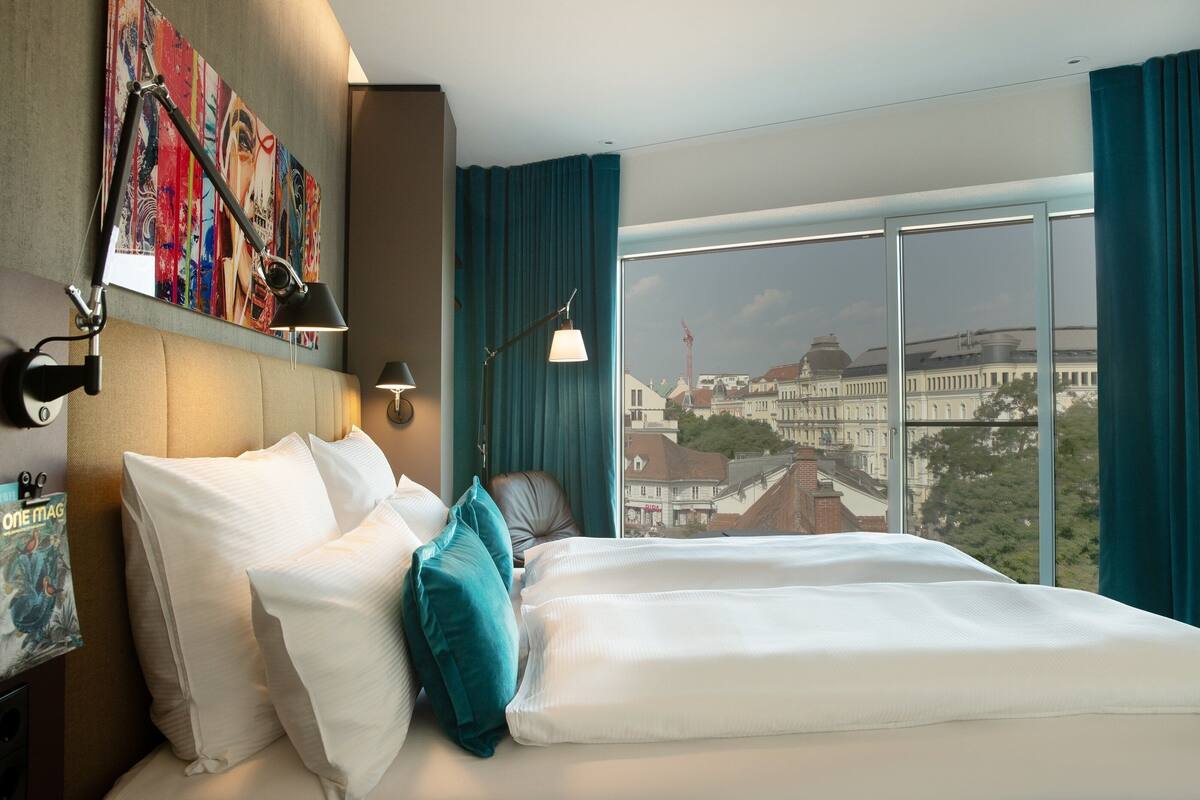 A hotel room with a large bed, a headboard with artwork, a nightstand with a lamp, and a view of a cityscape through a large window.