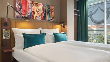 A hotel room with a large bed, a headboard with a cityscape painting, a wall-mounted lamp, and a window with a view of buildings.
