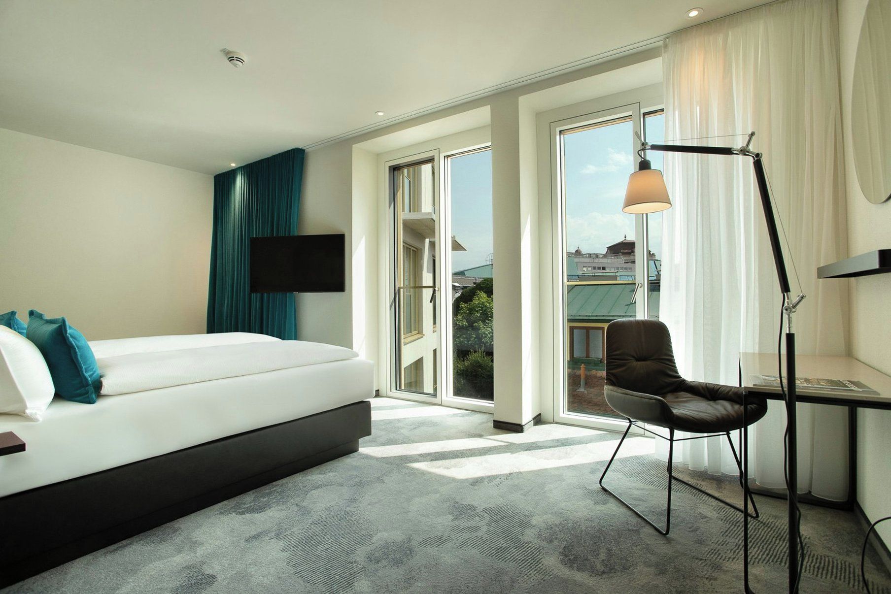 A hotel room with a bed, a desk, a chair, a TV, and a large window with a view of buildings.