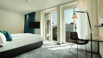 Habitación de hotel con cama, escritorio, silla, televisor y ventana grande con vista a edificios.