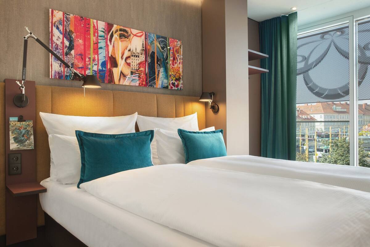 A hotel room with a large bed, a headboard with artwork, a nightstand with a lamp, and a view of the city through the window.