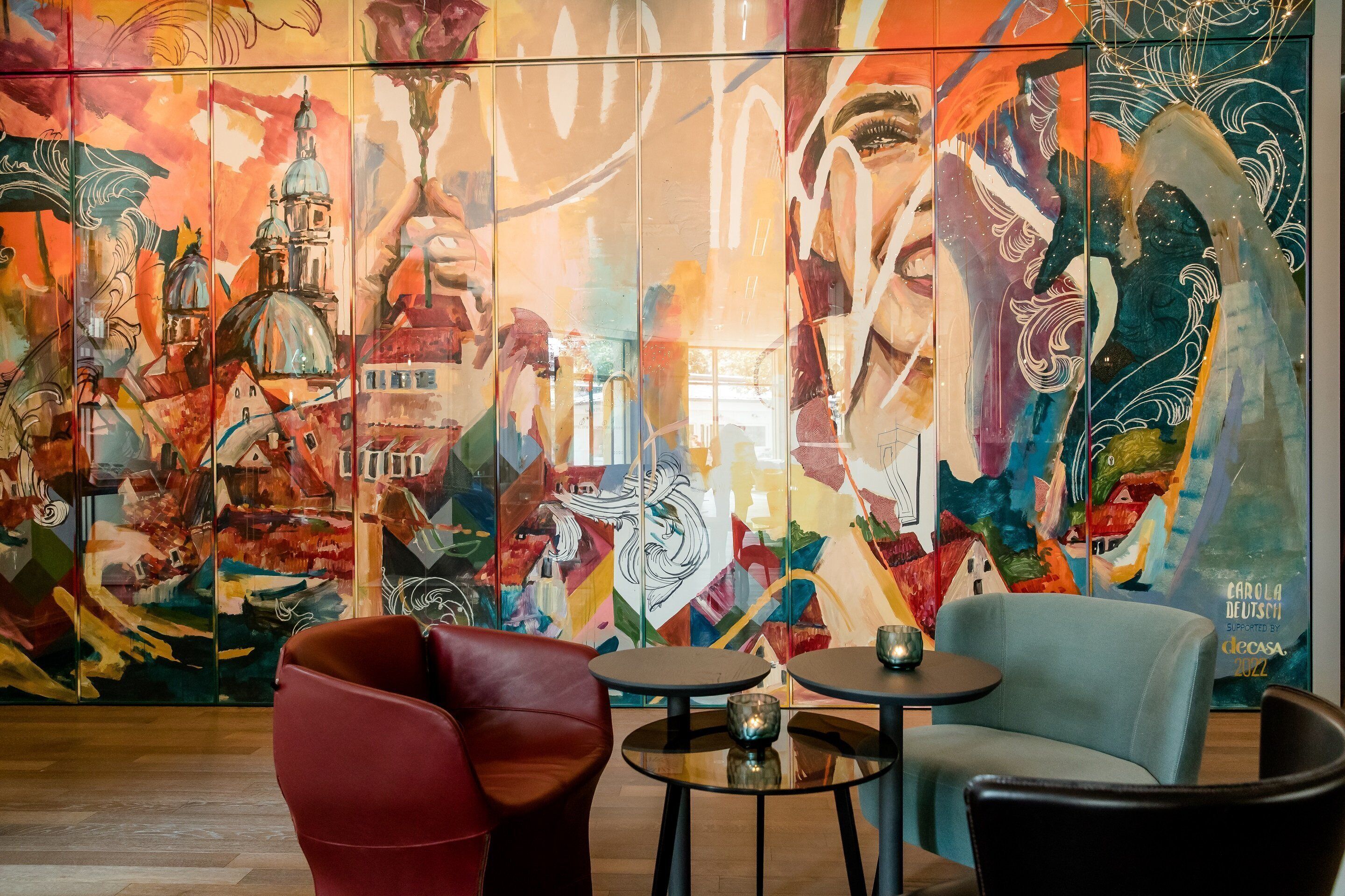A vibrant mural with abstract and architectural elements, a red chair, a blue chair, and two small round tables in a modern interior setting.