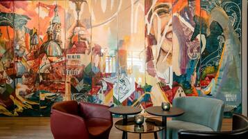 Un mural vibrante con elementos abstractos y arquitectónicos, una silla roja, una silla azul y dos pequeñas mesas redondas en un ambiente interior moderno.