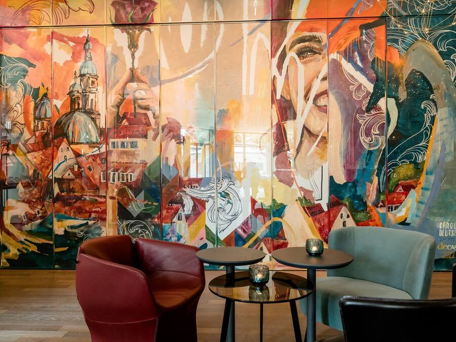 Un mural vibrante con elementos abstractos y arquitectónicos, una silla roja, una silla azul y dos pequeñas mesas redondas en un ambiente interior moderno.
