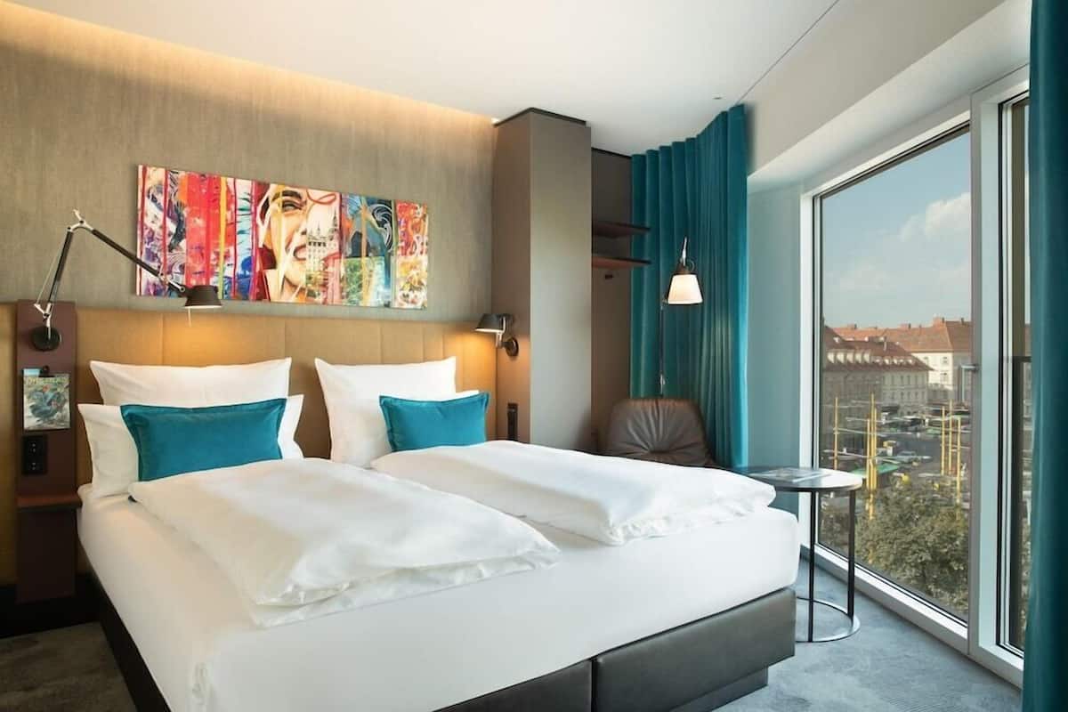 A modern hotel room with a large bed, a view of the city, and a painting on the wall.