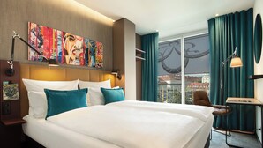 Ein Hotelzimmer mit einem großen Bett, einem Schreibtisch, einem Stuhl, einem Fenster mit Vorhängen und einem Bild an der Wand.