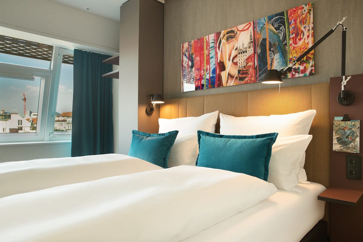 A hotel room with a large bed, teal pillows, a city view, and artwork on the wall.