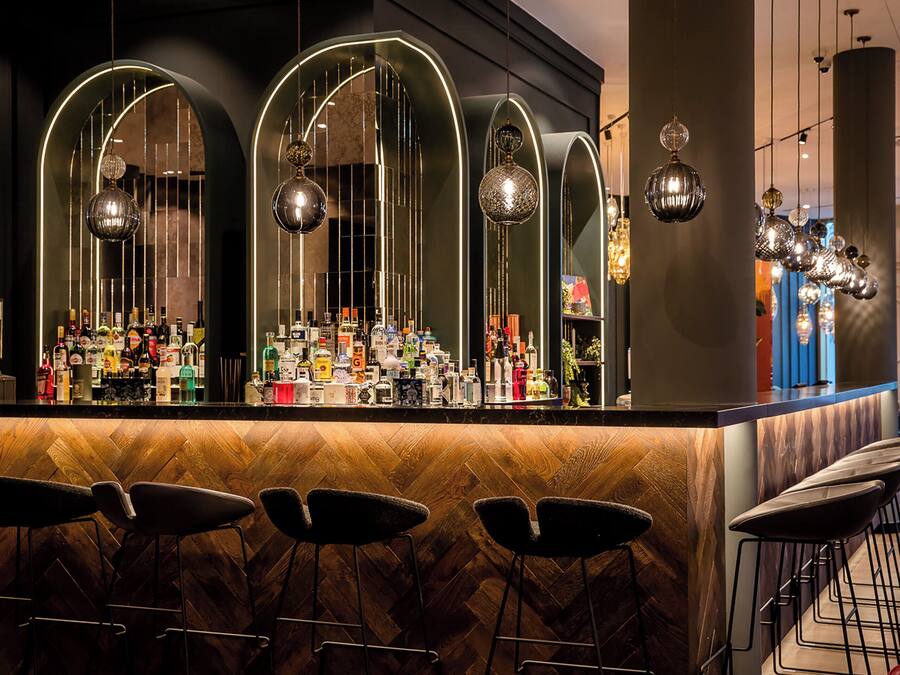 Uma área de bar com piso de madeira em padrão herringbone, banquetas e diversos copos e garrafas no balcão.