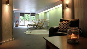 Een wellnessruimte met een massagetafel, een massagestoel, een klein tafeltje met een kaars en een raam met uitzicht op groen.