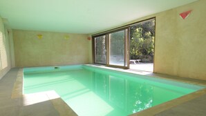 Une grande piscine intérieure aux parois en verre transparent, dotée d’un sol carrelé et offrant une vue sur les arbres à l’extérieur.
