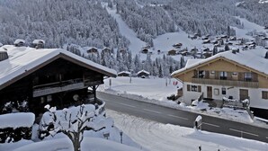 A snow-covered mountain village with wooden buildings, a road, and a ski slope. - GROTTE AUX FÉES A – Ski-in/ski-out apartment – ​​Wi-Fi – Parking (La Clusaz)