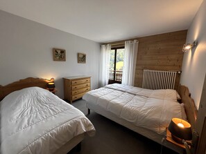 A bedroom with two beds, a wooden dresser, and a window with curtains. - GROTTE AUX FÉES A – Ski-in/ski-out apartment – ​​Wi-Fi – Parking (La Clusaz)