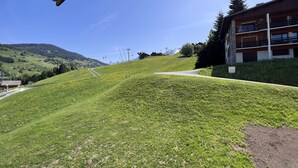 A grassy slope leading to a building with a balcony, surrounded by trees and a clear sky. - GROTTE AUX FÉES A – Ski-in/ski-out apartment – ​​Wi-Fi – Parking (La Clusaz)