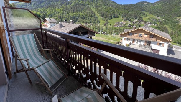 A balcony with a view of a mountain village, wooden buildings, and a clear sky. - GROTTE AUX FÉES A – Ski-in/ski-out apartment – ​​Wi-Fi – Parking (La Clusaz)