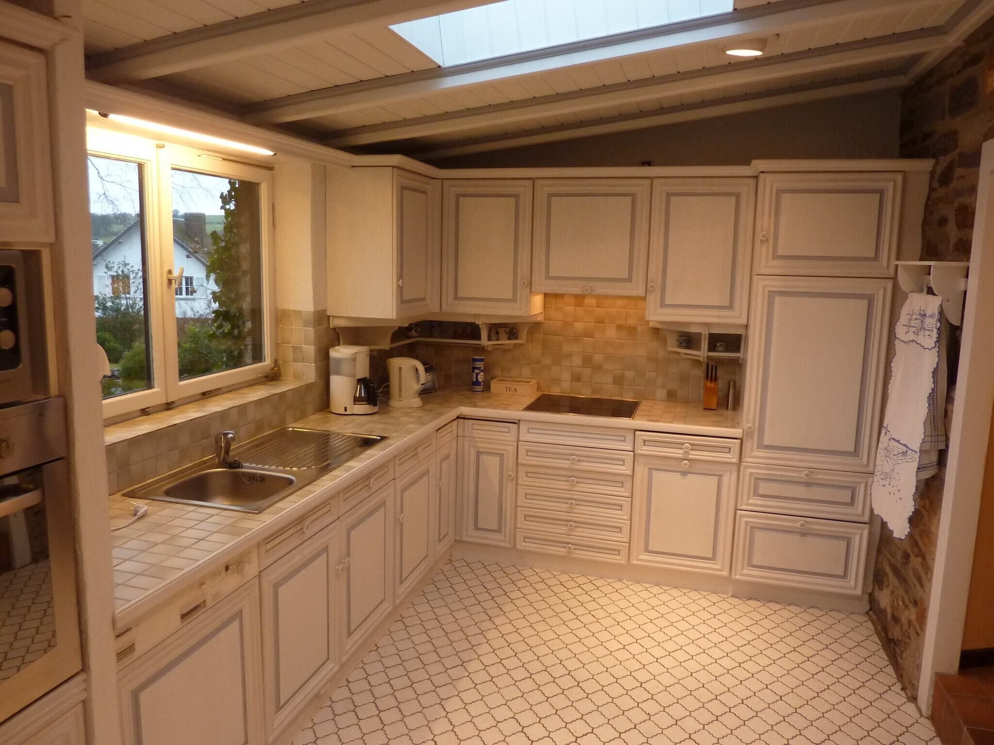 Cozinha com armários brancos, painel de azulejos, pia de aço inoxidável e janela com vista para uma casa.