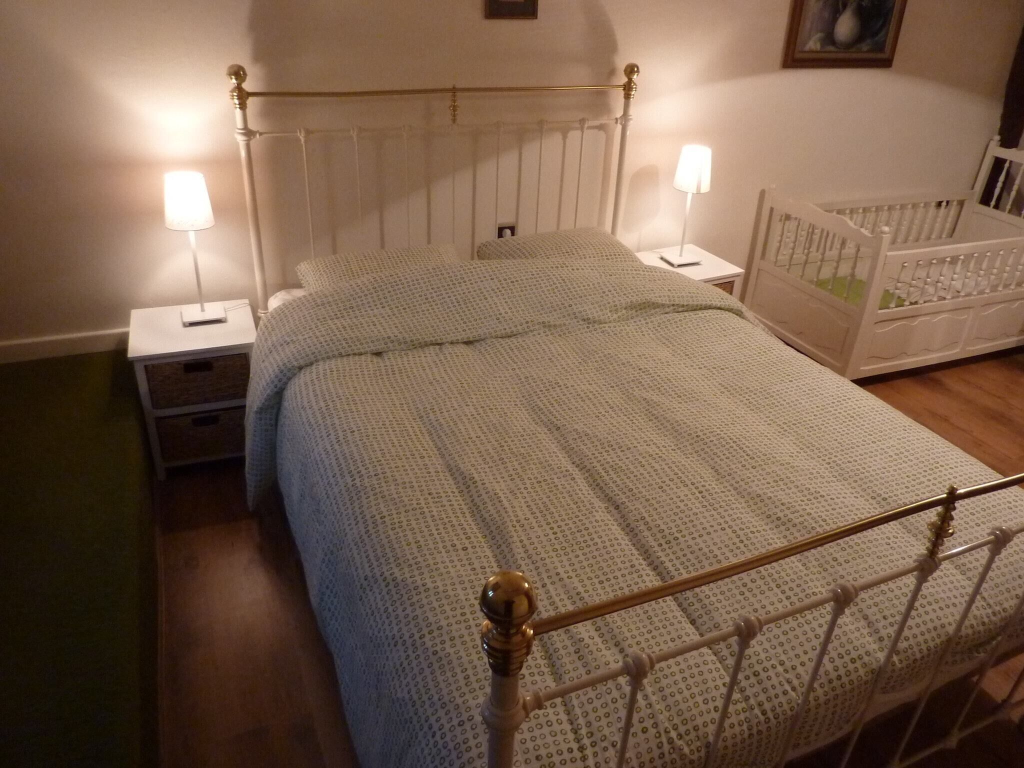 Ein ordentlich bezogenes Bett mit einem gemusterten Überwurf, zwei Nachttischlampen, ein Nachttisch und ein Babybett mit weißem Gestell.