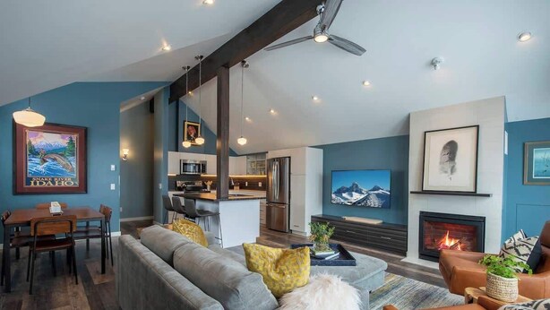 A modern living room with a sofa, coffee table, and fireplace. There is a kitchen in the background with a dining area.