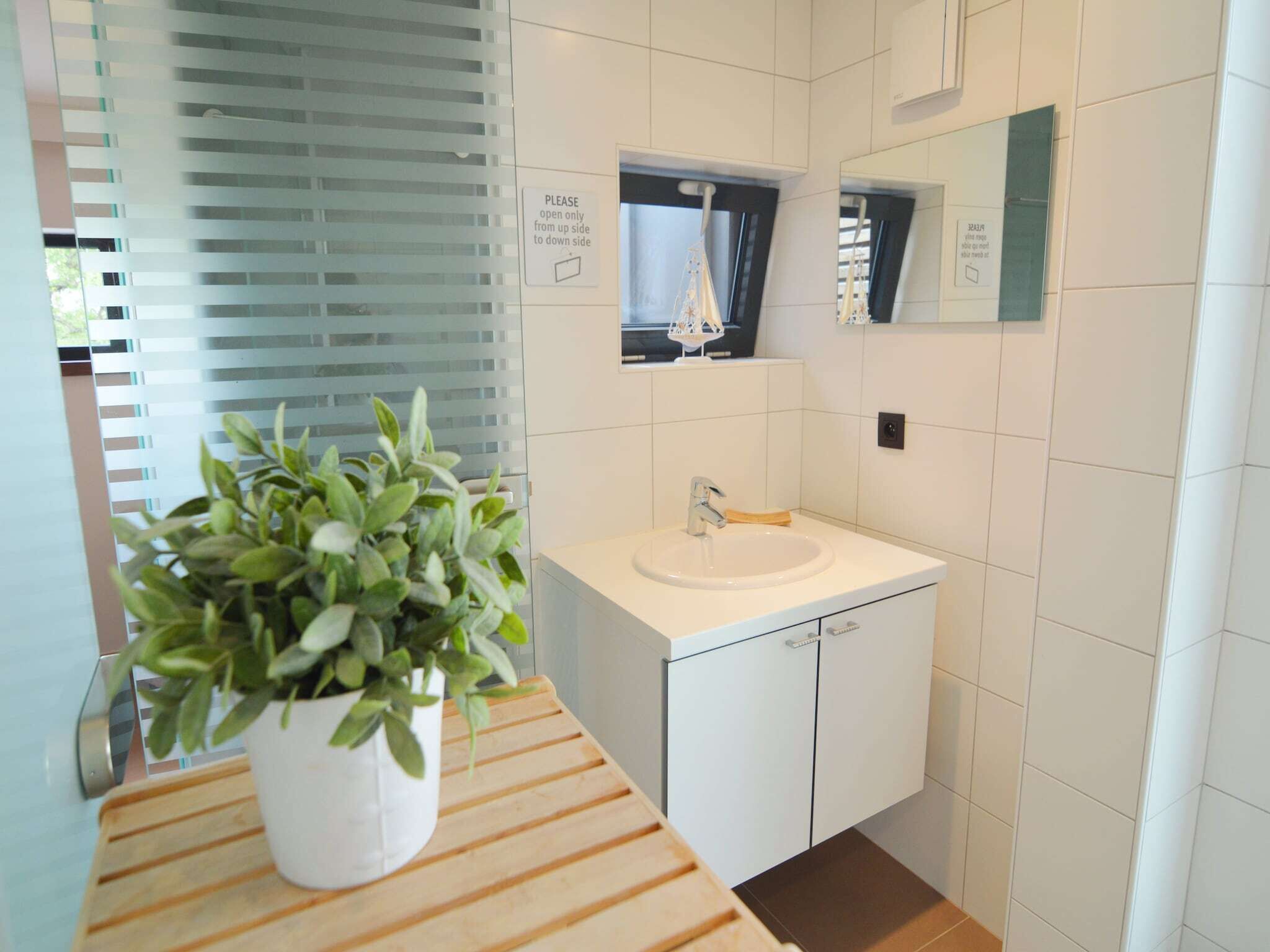 A modern bathroom with a white sink, a mirror, and a potted plant on a wooden shelf.
