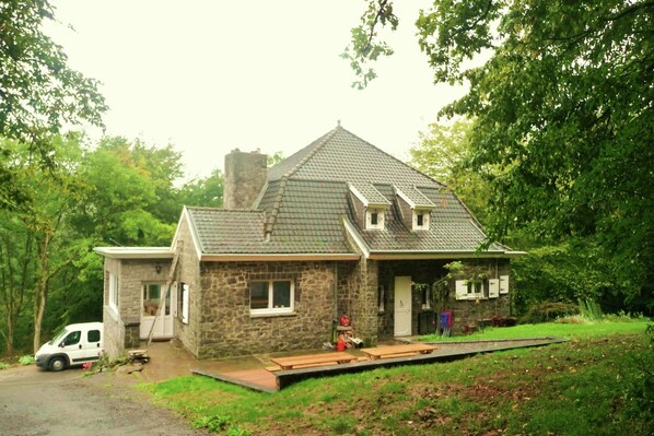 Een stenen huis met schuine dak en een klein terras, met een voertuig ervoor geparkeerd.