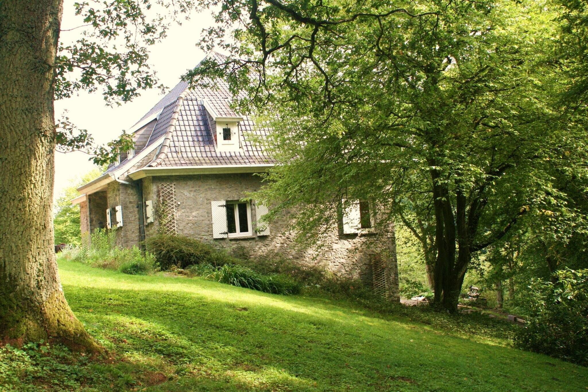 A stone house with a sloped roof, surrounded by lush greenery and trees.
