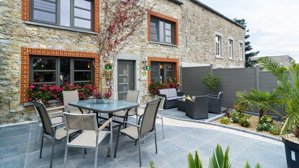 A stone building with a patio area featuring a round table and chairs, a small garden with flowers, and a grey fence. - Scenic Retreat in Gimnée (Gimnée)