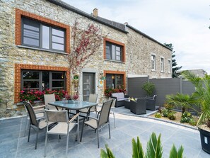 Un bâtiment en pierre doté d’un patio comprenant une table ronde et des chaises, un petit jardin fleuri et une clôture grise.