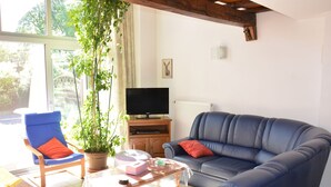 A living room with a blue sofa, a blue armchair, a glass coffee table, a potted plant, and a television on a wooden cabinet. - Nature Escape in Martelange (Martelange (radelange))