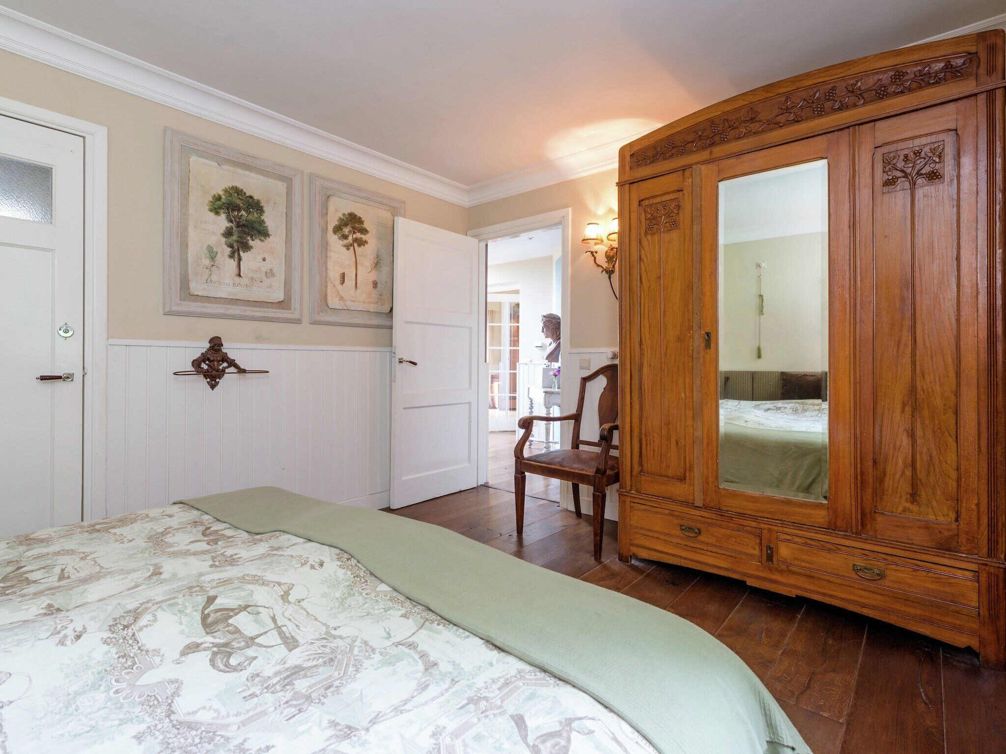 A bedroom with a large wooden wardrobe, a bed with a patterned bedspread, and a wall-mounted mirror.