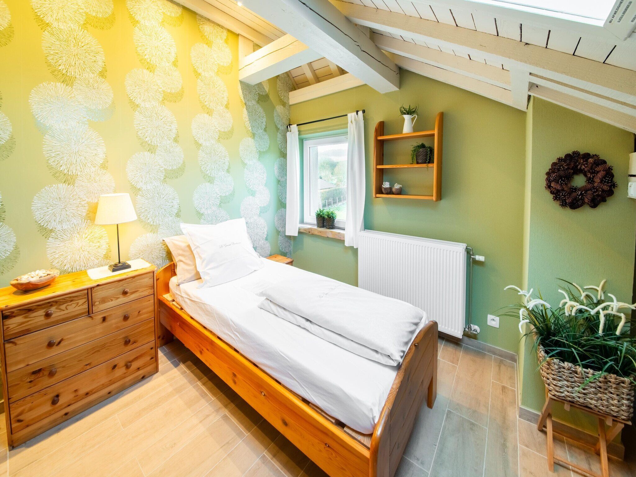 Une chambre à coucher avec un lit en bois, une table de chevet, une commode, une fenêtre donnant sur un beau paysage, un radiateur et une étagère avec des plantes.