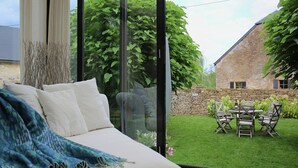 A modern living room with a sofa, a glass door leading to a garden with a stone wall and a table with chairs. - Rustic Charm, Modern Comfort (Latour)