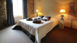 A bedroom with a stone wall, a bed with a white bedspread, two lamps, and a dress form on a stand. - Rustic Charm, Modern Comfort (Latour)
