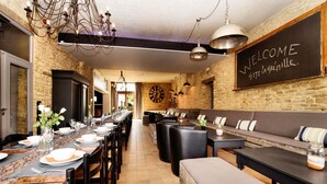 A restaurant with a long dining table set for a meal, a chalkboard with 'WELCOME' written on it, and a stone wall.