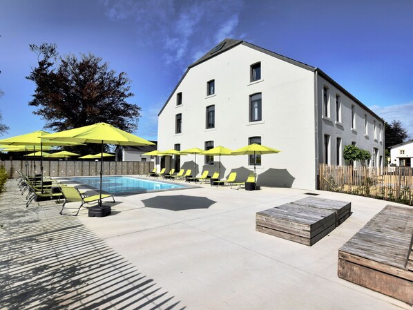A pool area with sun loungers and umbrellas, a wooden deck, and a white building with multiple windows.