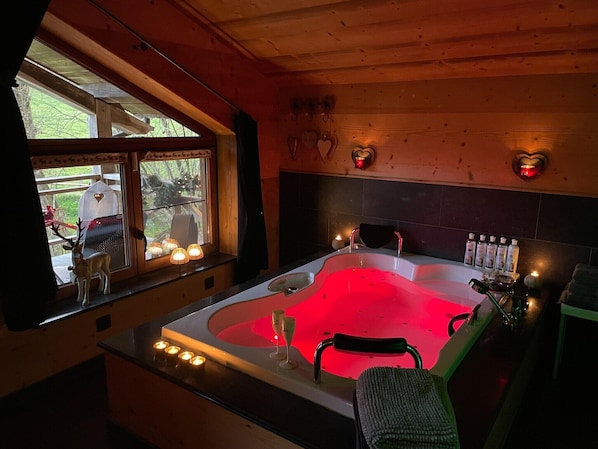 A bathroom with a large bathtub, a window with a view of the outside, and a shelf with toiletries.