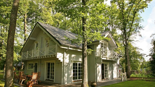 A two-story house with a porch, a dining area with chairs, and a large window. - Countryside Chalet in 	Bomal sur Ourthe with Sauna, Balcony (Bomal sur Ourthe)