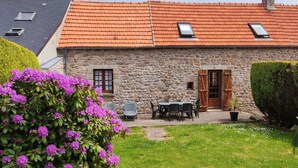 A stone house with a red-tiled roof, a small patio with outdoor seating, and a well-maintained garden with purple flowers.