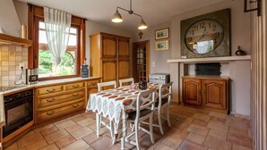 Une cuisine avec des meubles de rangement en bois, une table à manger avec des chaises, une grande horloge et une fenêtre avec des rideaux.