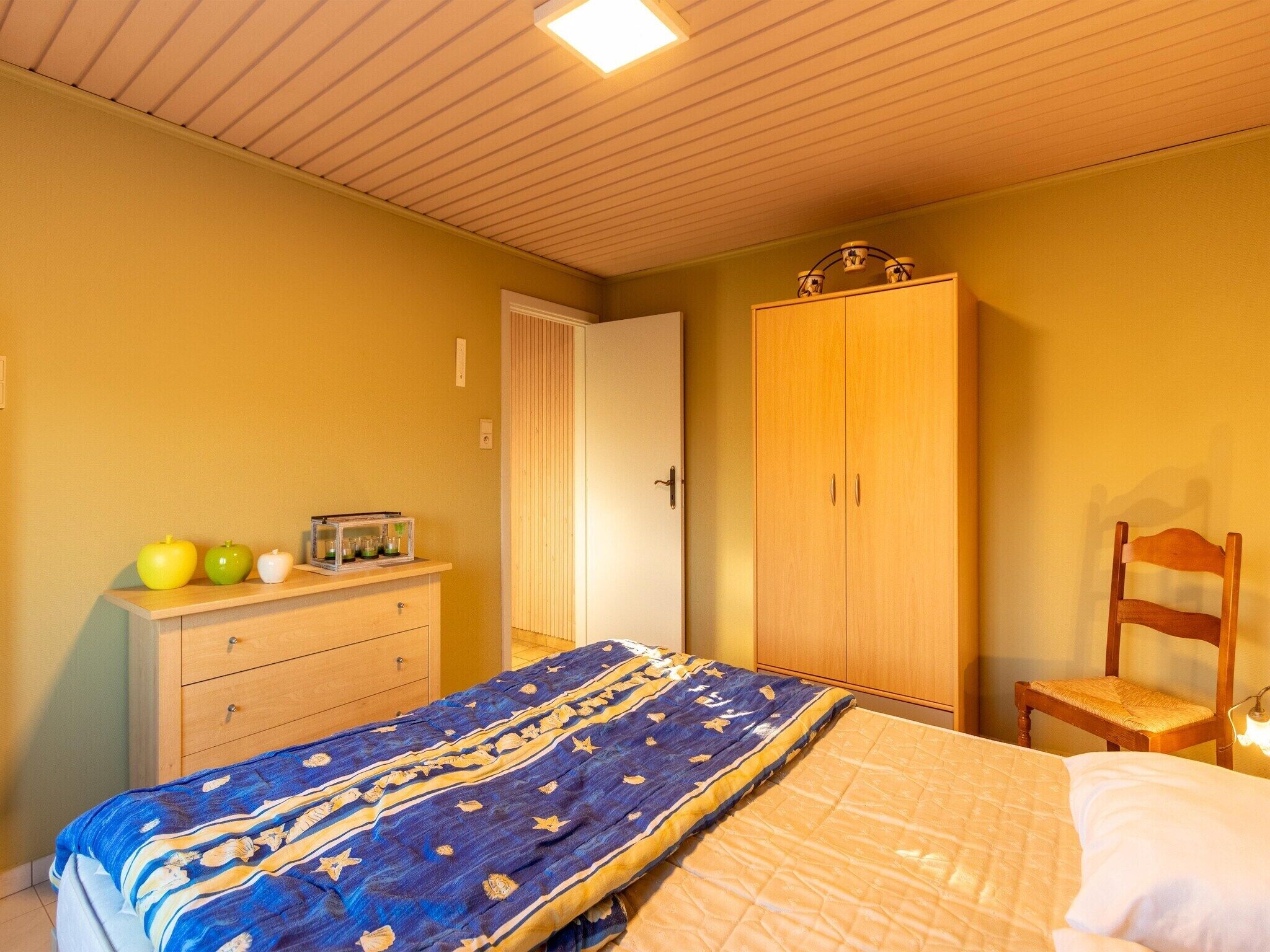 A bedroom with a bed, a wooden wardrobe, a chair, and a dresser with apples and a radio.