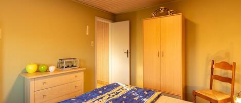 A bedroom with a bed, a wooden wardrobe, a chair, and a dresser with apples and a radio.
