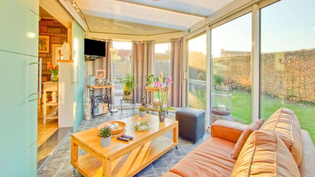 A modern living room with a wooden coffee table, a sofa, and a TV. There is a balcony with a view of the garden.
