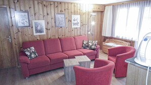 A cozy living room with a red sofa, a matching armchair, and a small wooden coffee table. There are framed pictures on the wooden wall, a lamp, and a window with curtains. - Chalet in Lenk near Simmen Falls (Lenk)