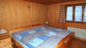 A wooden room with a bed, a nightstand, a lamp, a radiator, and a window with curtains.