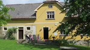 Un bâtiment jaune avec une façade en pierre, un escalier en pierre et un petit jardin agrémenté d’un banc et d’une piscine gonflable jaune.
