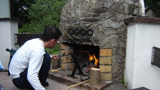 A stone-built outdoor oven with a fire burning inside, a person using a tool near the oven, and a wooden log on the ground. - Serene Holiday Home in Mladé Buky with small pool, Trampoline & Skiing Nearby (Mladé Buky)