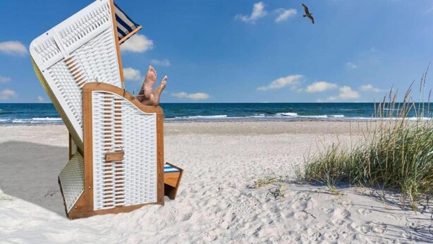 Une chaise de plage avec une structure en bois et un dossier en tissu blanc, placée sur une plage de sable, avec les vagues de l’océan en arrière-plan.