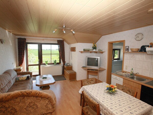 A cozy living room with a sofa, a dining table with chairs, a TV, and a kitchen area with a sink and shelves. - Ferienwohnung mit Terrasse in Kropelin (Kröpelin / OT Klein Nienhagen)